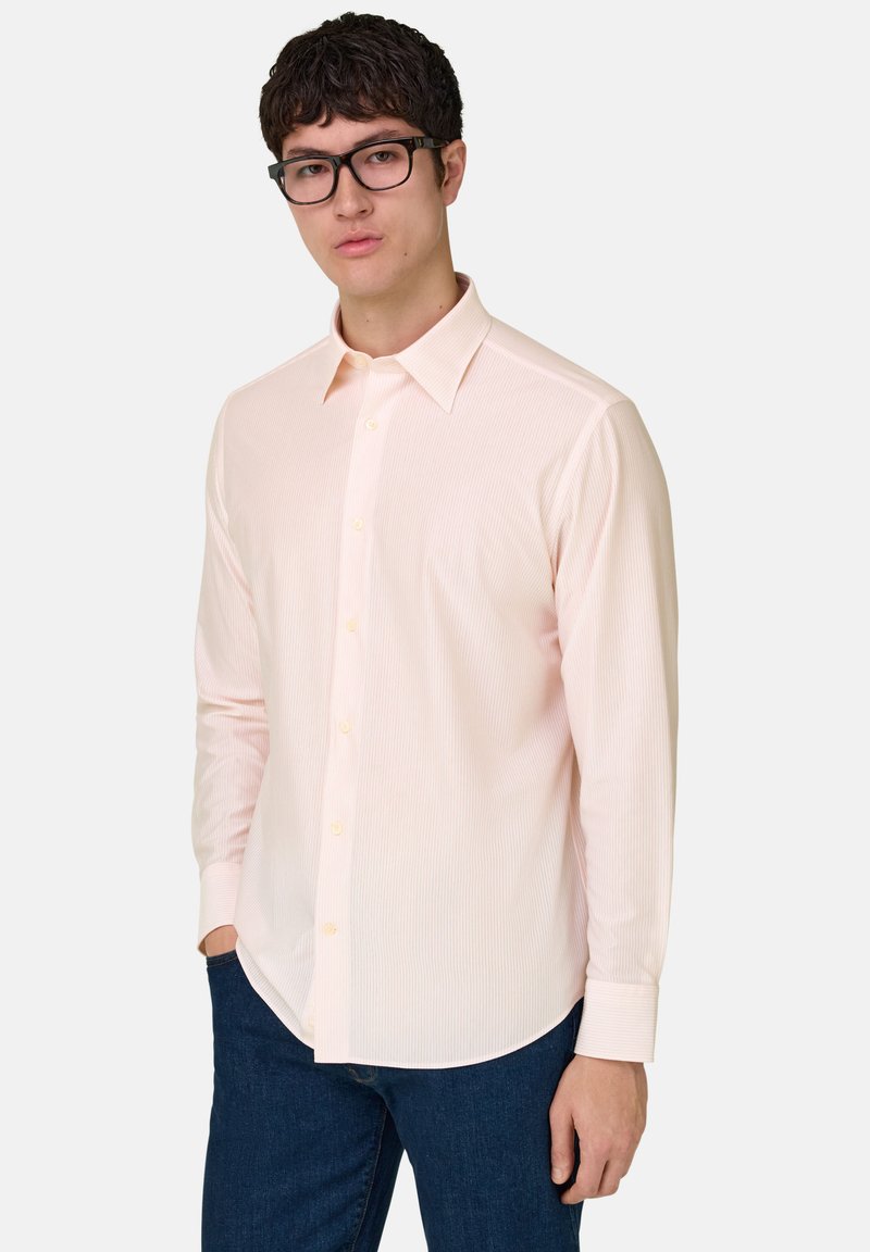Homme portant une chemise à boutons rose clair et un jean foncé, debout avec une main dans la poche devant un fond uni.