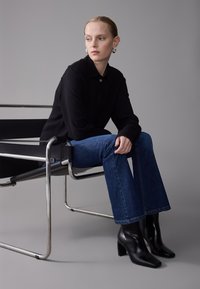Young woman with slicked-back hair wearing silver hoop earrings, black shirt, blue jeans, and black heeled boots sitting on a modern metal chair.