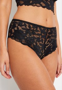 String en dentelle noire avec des motifs floraux, des bords festonnés et une texture semi-transparente. La taille est élastique pour plus de confort.