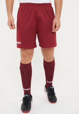 Homme portant un short de sport bordeaux, des chaussettes hautes assorties et des chaussures de football noires, debout sur un fond blanc.