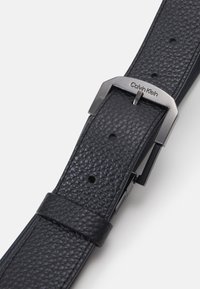 Cintura in pelle nera con superficie testurizzata, dotata di una fibbia in metallo color argento e logo Calvin Klein. Più fori per la regolazione.