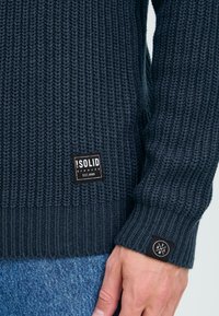 Knitted sweater in dark navy with ribbed texture. Features a black brand label on the chest and a round logo patch on the sleeve.