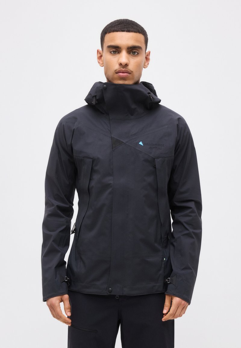 Νέος άνδρας που φοράει μαύρο αδιάβροχο μπουφάν με ψηλό γιακά, με φερμουάρ και κουμπιά, στέκεται μπροστά σε απλό ανοιχτόφωτο φόντο.