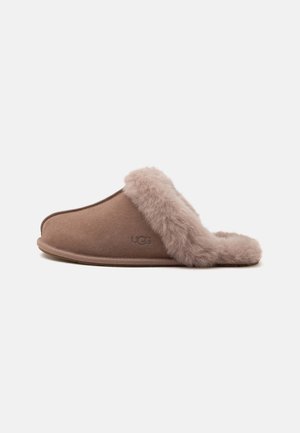 Καφέ παντόφλα τύπου slip-on με μαλακή, αφράτη επένδυση από γούνα και διακοσμητικά, με χαμηλό τακούνι και εξωτερικό υλικό που μοιάζει με σουέντ.