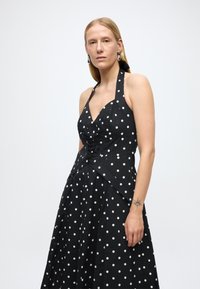 Vestido de tirantes negro con lunares blancos. Presenta un corpiño ajustado, detalles fruncidos y una falda en línea A. Tejido texturizado.