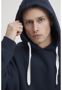 Námornícky modrá mikina s kapucňou vyrobená z mäkkej tkaniny, s veľkou kapucňou, bielymi šnúrkami s oranžovými akcentmi a voľným strihom.