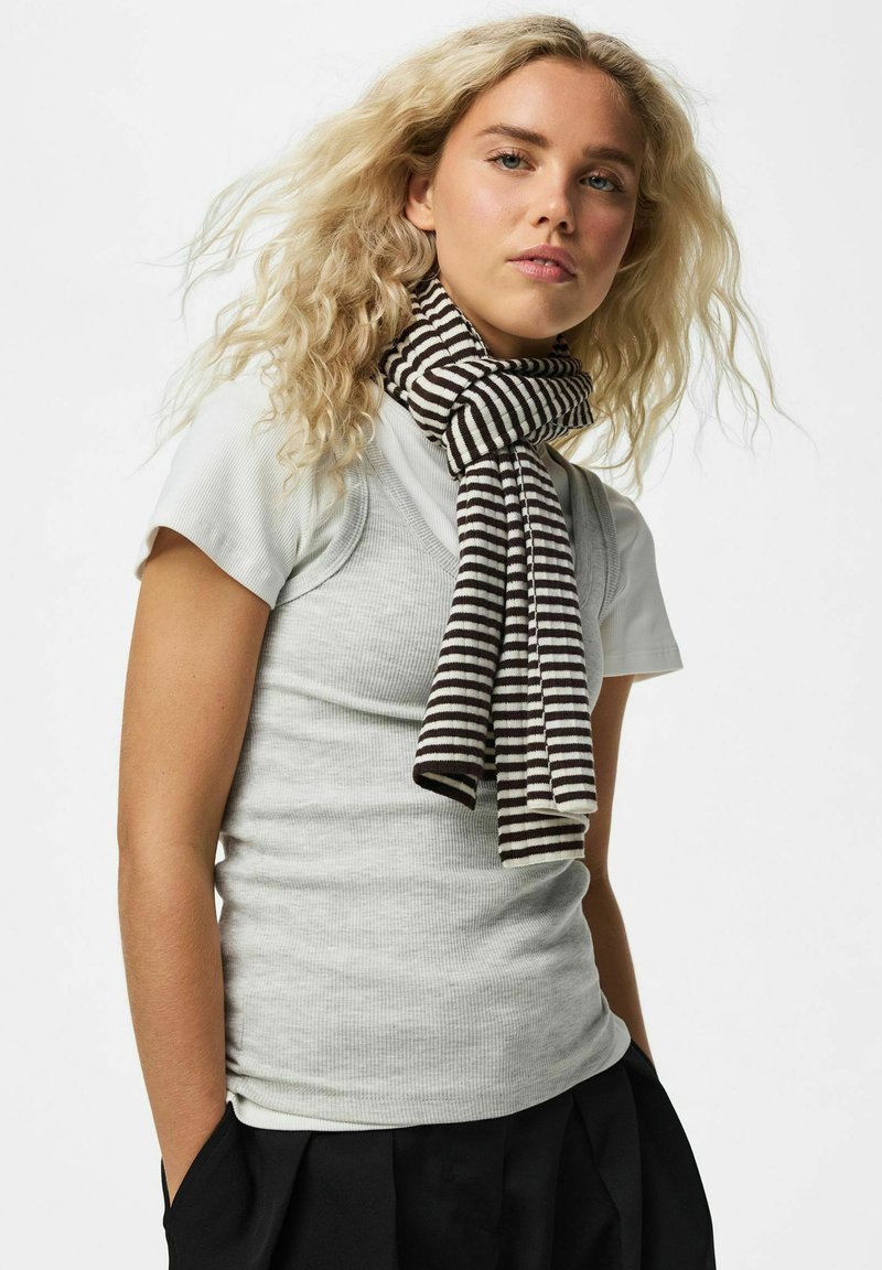 Jeune femme aux cheveux blonds bouclés porte un haut gris clair à manches courtes, un pantalon noir et une écharpe rayée noir et blanc.