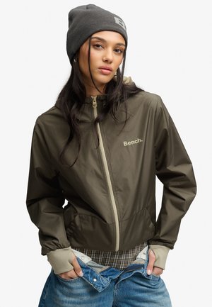 Jeune femme portant une veste Bench vert olive, un bonnet gris, une chemise à carreaux et un jean bleu avec les mains dans les poches du jean.