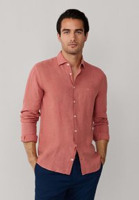 Mand står iført en koral pink skjorte med lange ærmer og knappet op, manchetterne er rullet op, samt marineblå bukser, hænderne i lommerne.