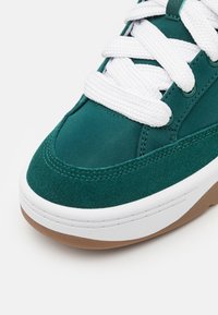 Vihreät mokka- ja kangaskengät, joissa on valkoinen nauhatoiminto, kumipohja ja litteä kärki, koristeena ommellut yksityiskohdat.