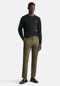 Schwarzer Sweatshirt mit Rundhalsausschnitt, kombiniert mit olivgrünen Hosen. Das Model trägt schwarze Schuhe, die einen Kontrast zum Outfit bilden.