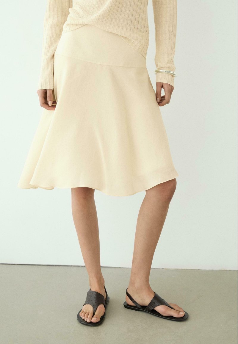 Vrouw draagt een crèmekleurige A-lijnrok, beige geribde lange mouwen top, zilveren armband en zwarte open-toe platte sandalen, staand op een grijze vloer.