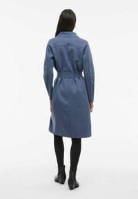 Cappotto blu a maniche lunghe con vita regolabile e dettaglio a plissè sul retro. Realizzato in tessuto morbido, presenta un colletto e una silhouette dritta.
