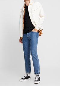 Hvid denimjakke over en orange plaid skjorte og sort t-shirt, parret med lyseblå jeans og sorte sneakers med hvide detaljer.