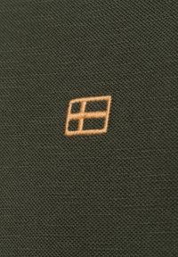 Ciemnozielony materiał o teksturowanym wykończeniu, z małym haftowanym pomarańczowym kwadratem w jednym rogu.