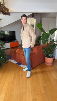 Chemise beige clair à boutons, jean bleu et baskets blanches. En arrière-plan, un canapé en velours côtelé marron, des plantes vertes et une unité TV.