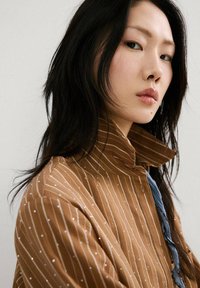 Mujer joven con cabello largo y negro, que lleva una chaqueta marrón a rayas, mirando ligeramente por encima del hombro sobre un fondo liso.