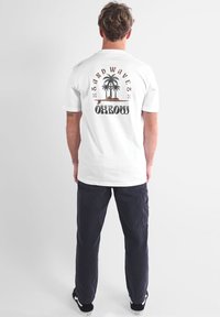 T-shirt en coton blanc avec un motif de palmier et le texte "SAND WAVES OXBOW" imprimé au dos. Associé à un pantalon bleu marine foncé et des baskets noires.