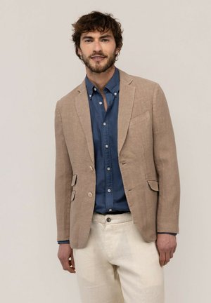Hombre con cabello rizado, vestido con blazer beige, camisa azul de botones y pantalones de colores claros, de pie frente a un fondo liso.