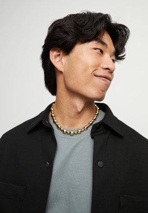 Joven con cabello negro que lleva una chaqueta negra, camisa gris y un collar grueso de cuentas doradas, mirando hacia la derecha con una sonrisa sutil.