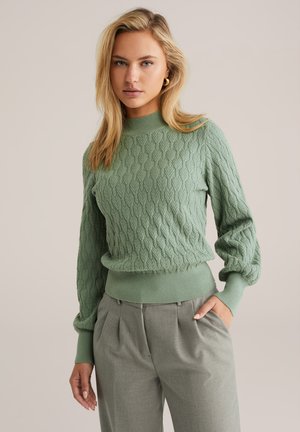 Pulover verde deschis din lână tricotată, cu gât înalt și mâneci evazate, asociat cu pantaloni eleganți gri deschis.