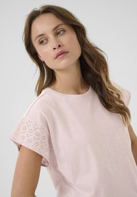 T-shirt rose clair à manches courtes. Les manches présentent des motifs floraux en œillets. Texture douce, encolure ronde, coupe décontractée.