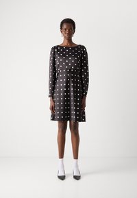 Abito nero con pois bianchi, maniche lunghe e scollatura rotonda. Tessuto liscio, forma svasata, lunghezza al ginocchio, abbinato a calze bianche e tacchi neri.