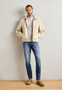 Jachetă ușoară bej, tricou gri, blugi albaștri și pantofi slip-on maro. Jacheta are manșete și talie elastice, având un croi lejer.