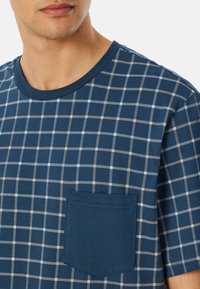 Blaues T-Shirt mit kurzen Ärmeln, das ein Karomuster in Beige und Weiß aufweist. Es hat eine aufgesetzte Tasche auf der linken Brust und einen runden Ausschnitt.