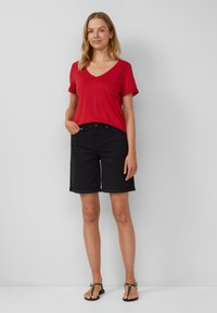T-shirt rouge à col en V et shorts en denim noir avec ourlets roulés. Le mannequin se tient les mains dans les poches, portant des sandales noires. Fond uni.