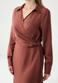 Sukienka kopertowa w rdzawym kolorze z kołnierzykiem i długimi rękawami, z cinchowanym wcięciem w talii i gładką teksturą.