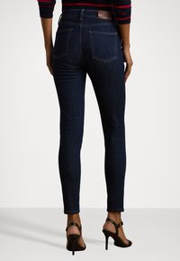 Skinny dark blue jeans with a smooth texture, high waistband, and rear pockets. Paired with black high-heeled sandals.