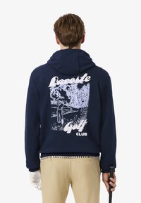 Felpa navy con cappuccio e coulisse, presenta una stampa grafica sul retro che ritrae un golfista. Orlo a coste con motivo a righe. Realizzata in tessuto morbido.