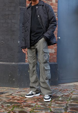 Persoon die buiten staat en een zwarte doorgestikte jas, zwart shirt, grijze cargobroek en zwart-witte sneakers draagt op een stenen bestrating.