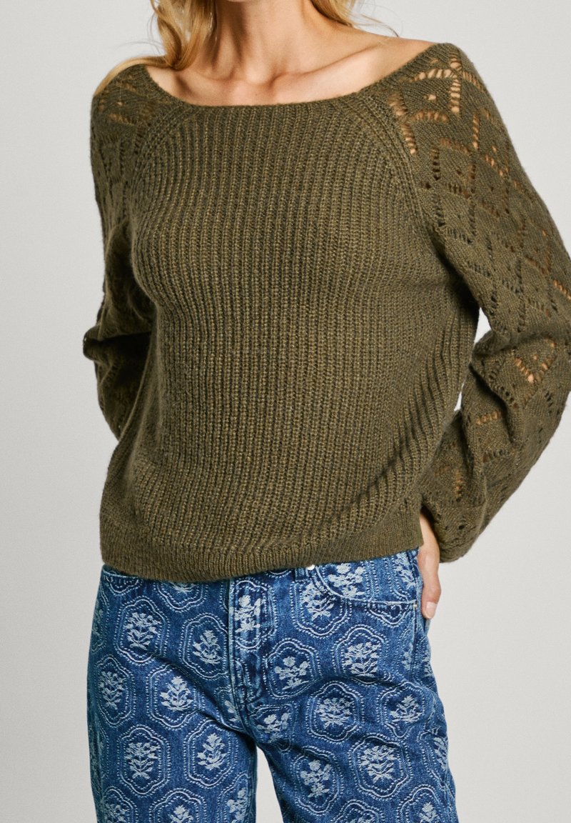 Olivgrüner gestrickter Pullover mit schulterfreiem Design, ausgestattet mit strukturierten Mustern an den Ärmeln und einem gerippten Saum. Kombiniert mit bedruckten blauen Jeans.