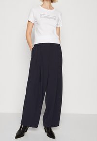 Valkoinen lyhythihainen t-paita, jossa on musta logopainatus, yhdistettynä korkeavyötäisiiin, poimutettuihin tummansinihin väljiin housuihin ja mustiin korkokenkiin.