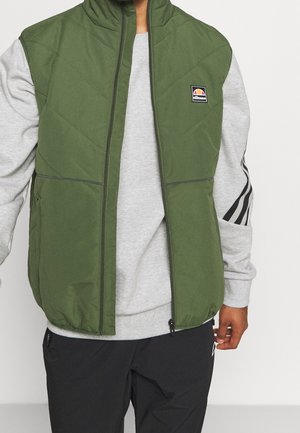 Gilet matelassé vert avec col montant et fermeture éclair. Dispose de deux poches avant et d'un petit logo sur la zone de la poitrine gauche. Superposé sur un sweat-shirt gris.