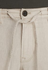 Pantalones de lino beige con cordón y cierre de botón de metal. Detalles de costura visibles, tejido texturizado y bolsillos laterales.