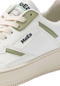 Białe skórzane sneakersy z zielonymi akcentami, sznurowana konstrukcja, teksturowana podeszwa oraz metka z logo "MoEa" z boku.