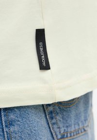Light beige cotton t-shirt with stitched hem, featuring a black branded label on the side, paired with blue denim jeans.