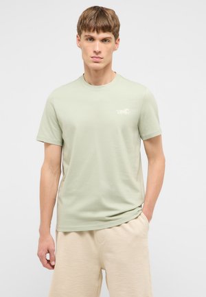 Junger Mann trägt ein hellgrünes T-Shirt mit kurzen Ärmeln und kleinem weißem Logo sowie beigefarbene Shorts, steht vor einfarbigem Hintergrund.