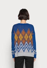 Stickad tröja i blått, med diamantmönster i orange och gult, samt en grå nederkant. Mjuk textur och oversized passform.