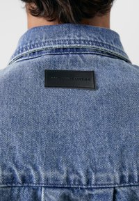 Jaqueta de denim azul claro com um rótulo de couro preto na parte de trás, com texto em relevo que diz "NÃO DESPERDICES CULTURA". Gola clássica e costura.