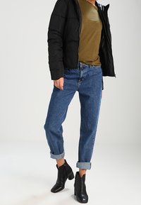 Giubbotto di piuma nero, t-shirt verde oliva, jeans blu arrotolati, e stivaletti neri con una texture liscia e punta appuntita.