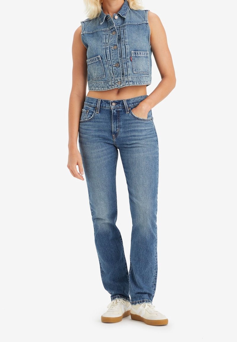 Femme portant une veste en jean courte sans manches bleu clair et un jean taille haute droit avec des baskets blanches, debout avec une main dans la poche.