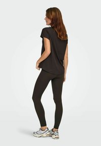 Černý top s krátkým rukávem a volným střihem, v kombinaci s tmavými žebrovanými legínami. Modelka má na sobě stříbrné sportovní boty, pohled zezadu.
