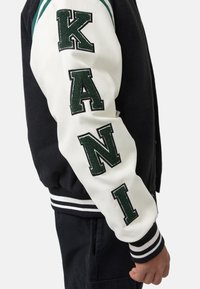 Jachetă varsity alb-negru cu litere verzi brodate "KANI" pe mânecă, cu manșete rigide cu dungi și accente pe umeri.