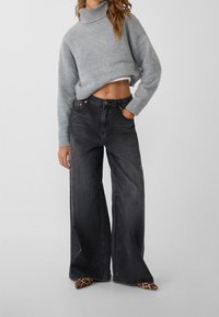 Grijze, geknipte trui met een hoge kraag, gecombineerd met zwarte, wijde jeans met een knoop en zakken. Schoenen met een luipaardprint.