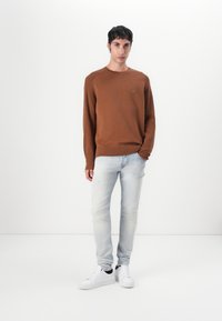 Rjava pletena majica s krožnim izrezom, uparjena s svetlo modrimi jeans hlačami in belimi supergami. Ima majhen logotip na prsih.