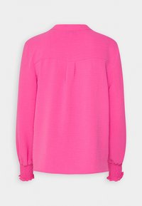 Blouse rose vif à manches longues vue de dos, avec poignets froncés et un pli sous l'empiècement. Pas de boutons ou de motifs visibles.